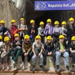 Grupa dzieci w żółtych kaskach siedzi razem w kopalni soli pod dużym znakiem.