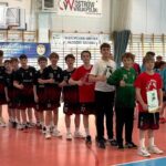 Drużyny piłki ręcznej chłopców ustawiają się na krytym boisku, niektóre trzymają trofea i certyfikaty.