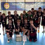 Chłopięca drużyna sportowa świętuje w pomieszczeniu z medalami i trofeum, pozując przed banerem.