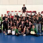 Młodzieżowa drużyna piłki ręcznej świętuje z trenerami, medalami i certyfikatami przed bramką.