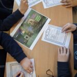 Czterech uczniów pisze na arkuszach wokół stołu z kolorowym arkuszem przedstawiającym robota w centrum.