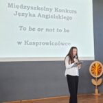 Kobieta przemawia na scenie w pobliżu ekranu wyświetlającego tekst Międzyszkolnego Konkursu Języka Angielskiego.