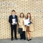 Trzy osoby trzymające certyfikaty stoją przed ceglaną ścianą z polskim tekstem.