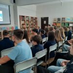 Uczniowie siedzą w bibliotece, oglądając prezentację z obrazami wyświetlanymi na ekranie z przodu.