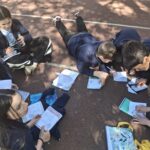 Dzieci siedzą i leżą na ziemi na świeżym powietrzu, pisząc w zeszytach i pracując razem w grupach.