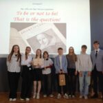 Ośmiu uczniów stoi na scenie, a za nimi wyświetlana jest prezentacja Hamleta o Szekspirze.