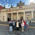 Grupa dzieci pozuje przed zabytkowym budynkiem z flagami i ozdobnymi dekoracjami w słoneczny dzień.