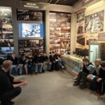 Grupa dzieci siedzi na wystawie muzealnej, słuchając przewodnika w pobliżu historycznych zdjęć i artefaktów.