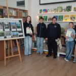 Sześciu uczniów stoi przy tablicy i półce z książkami, trzymając certyfikaty w klasie lub bibliotece.