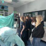 Grupa uczniów obserwuje demonstrację procedury medycznej obok przykrytego pacjenta w sali szpitalnej.