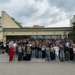 Duża grupa uczniów i nauczycieli pozuje przed budynkiem szkoły w pochmurny dzień.