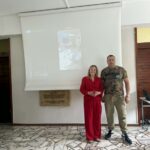 Kobieta w czerwonym garniturze i mężczyzna w kamuflażu stoją przed pustym ekranem projektora.