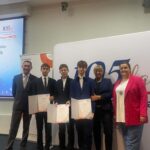 Pięć osób w oficjalnych strojach trzyma certyfikaty i pozuje podczas ceremonii wręczenia nagród, mając za sobą banery.