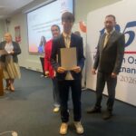 Młody mężczyzna trzymający certyfikat podczas ceremonii wręczenia nagród, z trzema dorosłymi stojącymi w pobliżu.