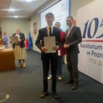 Młody mężczyzna w garniturze trzyma certyfikat podczas ceremonii wręczenia nagród w pomieszczeniach zamkniętych.