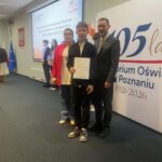 Trzy osoby pozują w pomieszczeniu z certyfikatem; inni stoją w kolejce w pobliżu ekranu prezentacji.