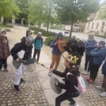 Dwójka dzieci bawi się na zewnątrz tarczami i rekwizytami, obserwowana przez grupę dzieci i dorosłych.