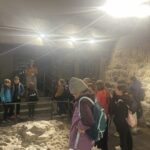 Grupa dzieci z plecakami ogląda wystawę archeologiczną z kamieniami i piaskiem.