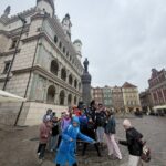 Grupa ludzi pozuje przed zabytkowym budynkiem i pomnikiem na europejskim rynku.
