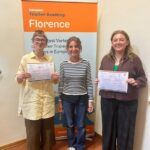 Dwie osoby trzymają certyfikaty, stojąc z inną osobą przed znakiem Florence Teacher Academy.
