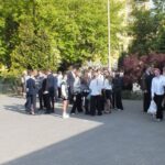 Grupy uczniów w formalnych strojach zbierają się na zewnątrz w słoneczny dzień w pobliżu drzew i krzewów.