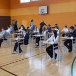 Uczniowie siedzą przy rozstawionych ławkach w sali gimnastycznej, przygotowując się do egzaminu.