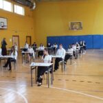 Uczniowie siedzą przy pojedynczych biurkach w sali gimnastycznej, rozstawieni w odstępach, przygotowując się do egzaminu.
