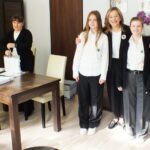 Dyrektor i nauczyciele w formalnych czarno-białych strojach w jasnym, nowoczesnym pomieszczeniu ze stołami.