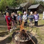 Dzieci pieką kiełbaski nad ogniskiem na zewnątrz drewnianego budynku.
