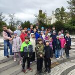 Grupa dzieci w kolorowych kurtkach pozuje razem na zewnętrznych schodach przed drzewami i budynkiem.