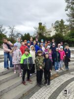 Grupa dzieci w kolorowych kurtkach pozuje razem na zewnętrznych schodach przed drzewami i budynkiem.