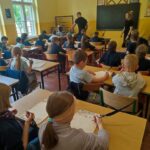 Dzieci siedzą w ławkach w klasie, pisząc na tablicach, podczas gdy nauczyciele stoją z przodu.