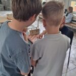 Dwóch chłopców stoi blisko siebie, grając w grę stołową w jasnej klasie z dużymi oknami.