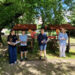 Grupa ludzi stoi na zewnątrz pod drzewami w pobliżu stołów piknikowych i kolorowych lampionów w słońcu.