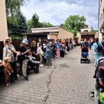 Grupa ludzi, w tym dzieci, gromadzi się na zewnątrz wzdłuż utwardzonego chodnika, między budynkami a drzewami.