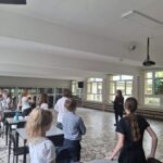 Nauczyciel stoi z przodu klasy, a uczniowie stoją przy swoich ławkach, zwróceni w jej stronę.