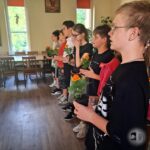 Grupa dzieci stoi w szeregu w pomieszczeniu, a każde z nich trzyma w rękach małą doniczkę z nagietkiem.