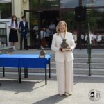 Kobieta w białym garniturze trzyma trofeum, stojąc na zewnątrz budynku w pobliżu stołu z innym trofeum.