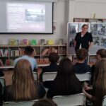 Kobieta prowadzi prezentację dla uczniów w sali lekcyjnej wyposażonej w regały z książkami i ekran projekcyjny.