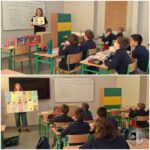 Nauczyciel pokazuje kolorową tablicę skupionym uczniom w klasie, w której na ławkach leżą książki.