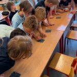 Dzieci pochylają się nad ławkami w klasie, przyglądając się małym prostokątnym przedmiotom podczas zajęć grupowych.