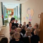 Nauczyciel rozmawia z grupą młodych uczniów na szkolnym korytarzu, tuż obok ściennej malowidła przedstawiającego postacie z kreskówek.