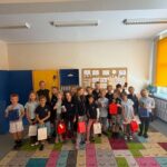 W kolorowej sali lekcyjnej stoi grupa uśmiechniętych dzieci trzymających torby z prezentami.