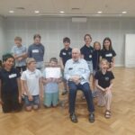 W jasnej sali lekcyjnej siedzi i stoi grupa uśmiechniętych dzieci wraz ze starszym mężczyzną.