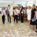Uczniowie w strojach uroczystych niosą flagę wzdłuż przejścia, podczas gdy pozostali stoją i obserwują to w szkolnej auli.