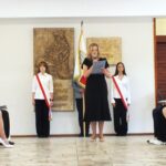 Kobieta przemawia z podium w pomieszczeniu, po bokach stoją uczniowie trzymający flagi, a inni siedzą w pobliżu.