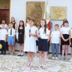 Grupa uczniów stoi w pomieszczeniu; dwie dziewczynki z przodu trzymają mikrofony, reszta stoi z tyłu.