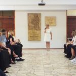 W jasnym korytarzu wewnątrz budynku stoi kobieta ubrana na biało i przemawia do dwóch rzędów siedzących osób.