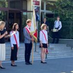 Uczniowie w mundurkach stoją z flagą podczas szkolnej uroczystości; za nimi, na schodach, stoi perkusista.