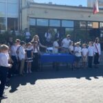 Grupa uczniów i nauczycieli stoi przed szkołą podczas uroczystości w słoneczny dzień.